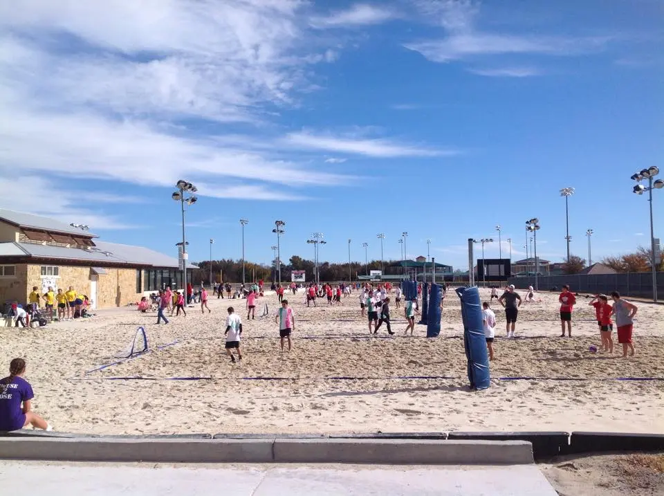 Sandy recreational area with people playing volleyball, tall lights, and facility buildings under blue sky