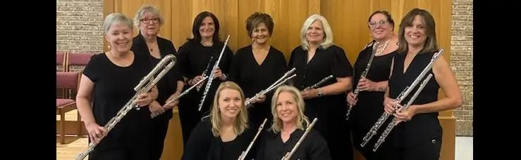 Nine women in black clothing holding flutes, posing together in a wooden-paneled room