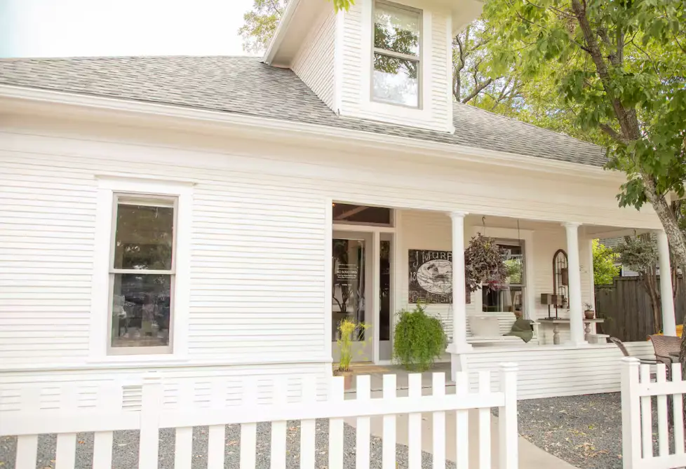White farmhouse with covered front porch, white picket railing, and potted plants flanking the entrance door