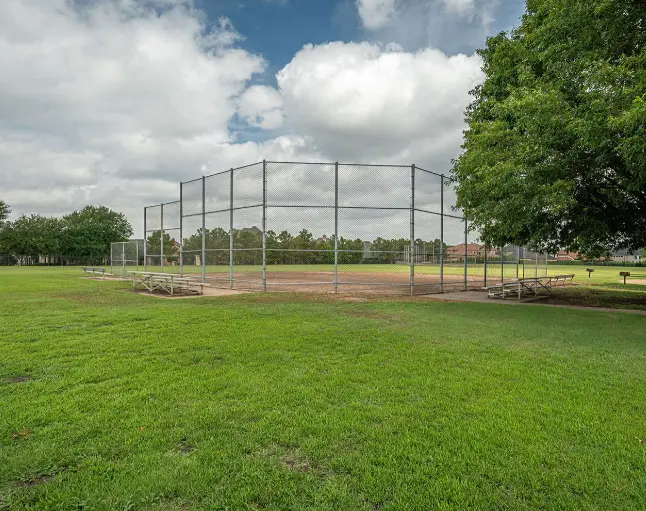Metal batting cage on green grass field with trees and blue sky with white clouds