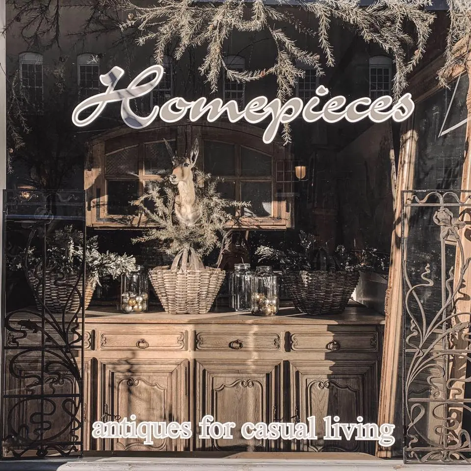 Storefront window display with dried botanicals in woven baskets and glass vases, vintage wooden cabinet beneath, ornate iron doors flanking entrance