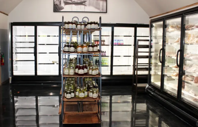 Glass-fronted refrigerated display cases surrounding a wooden shelving unit stocked with bottles and jars of condiments and preserved goods