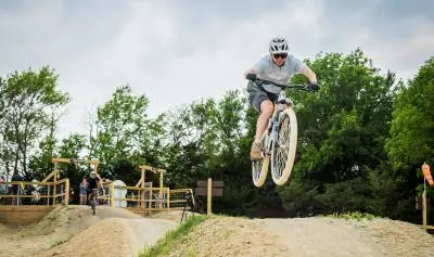 Mountain biker airborne on dirt jump trail with spectators and green trees in background