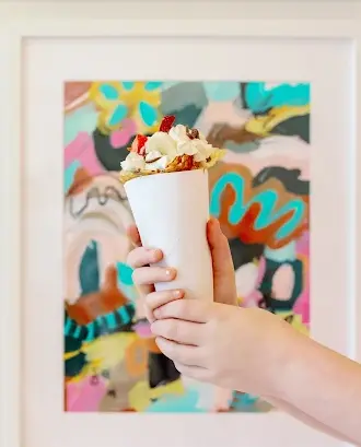Hand holding a white cup of crepe topped with whipped cream and granola against colorful abstract mural background