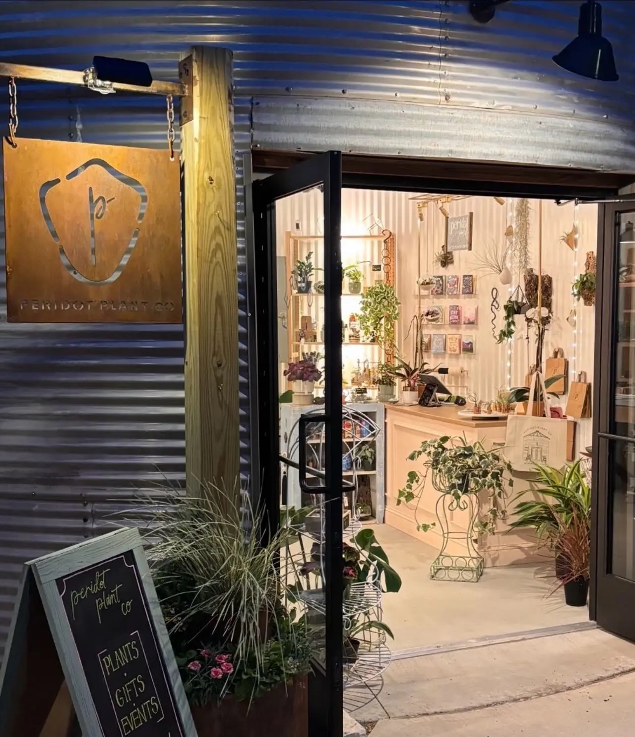Plant shop storefront with open doors, potted plants outside, and interior display of hanging plants, botanical artwork, and home décor visible through windows