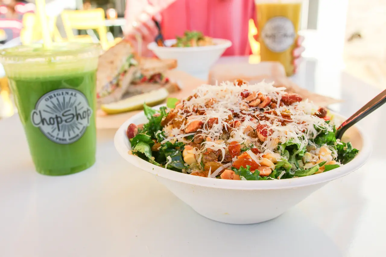 Bowl of chopped salad with greens, vegetables, and shredded cheese, alongside green juice in branded cup
