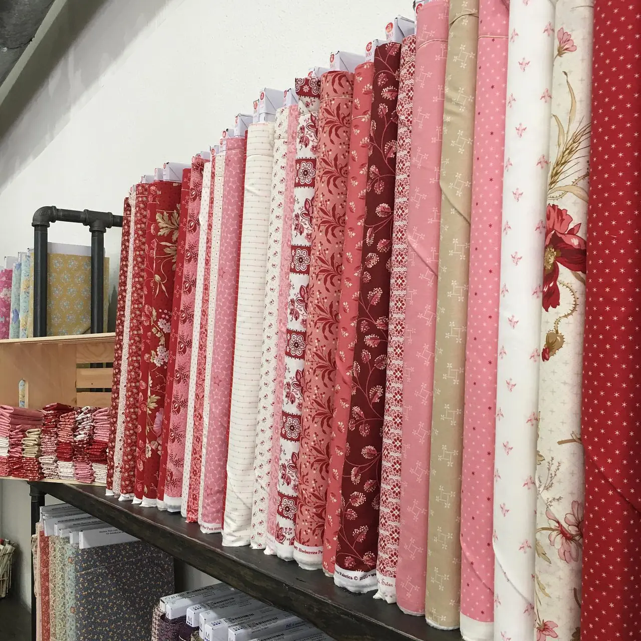 Bolts of fabric in shades of red, pink, cream and beige displayed on a black shelf, featuring various patterns including florals, checks and geometric designs