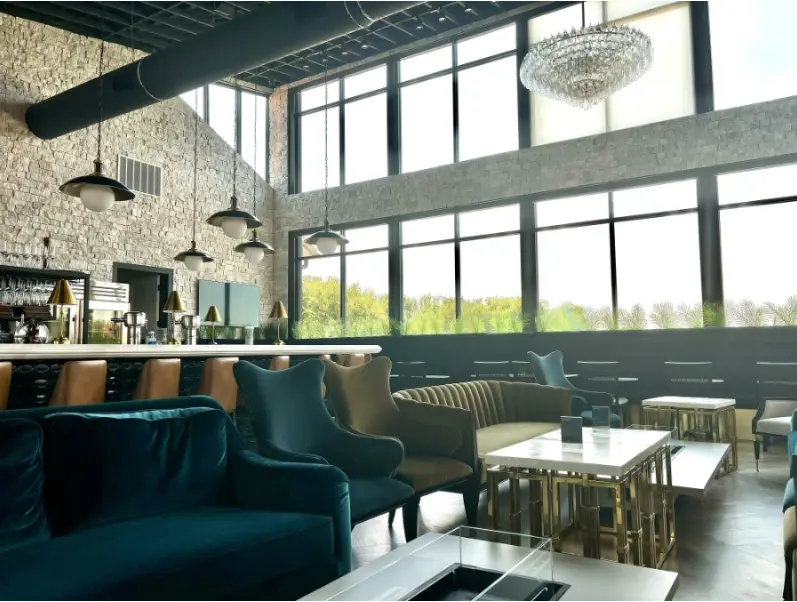 Modern lounge with teal velvet seating, exposed brick walls, floor-to-ceiling windows, and contemporary lighting fixtures overlooking greenery