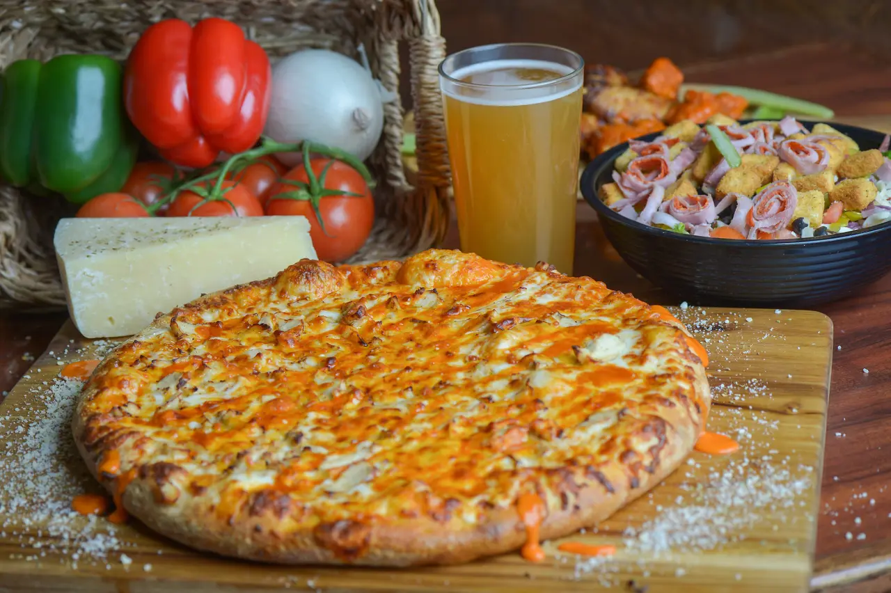 Thick-crust pizza with melted cheese on wooden board, fresh vegetables, cheese wedge, glass of beer, and pasta salad in background