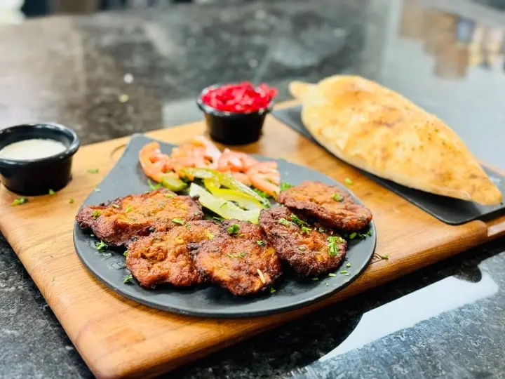 Grilled meat skewers with cucumber and tomato garnish, flatbread, and small bowls of sauce on a wooden serving board