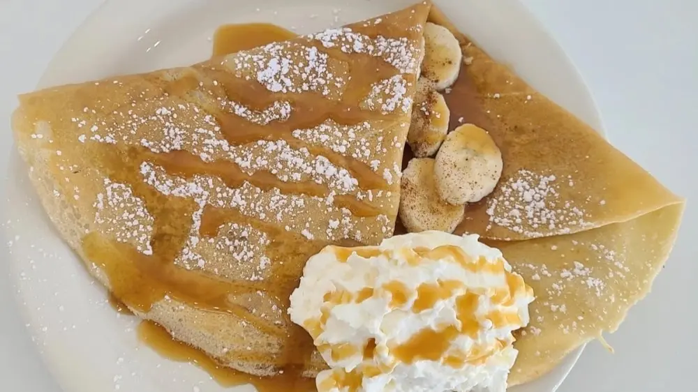 Folded crepes dusted with powdered sugar, topped with sliced bananas and whipped cream on a white plate