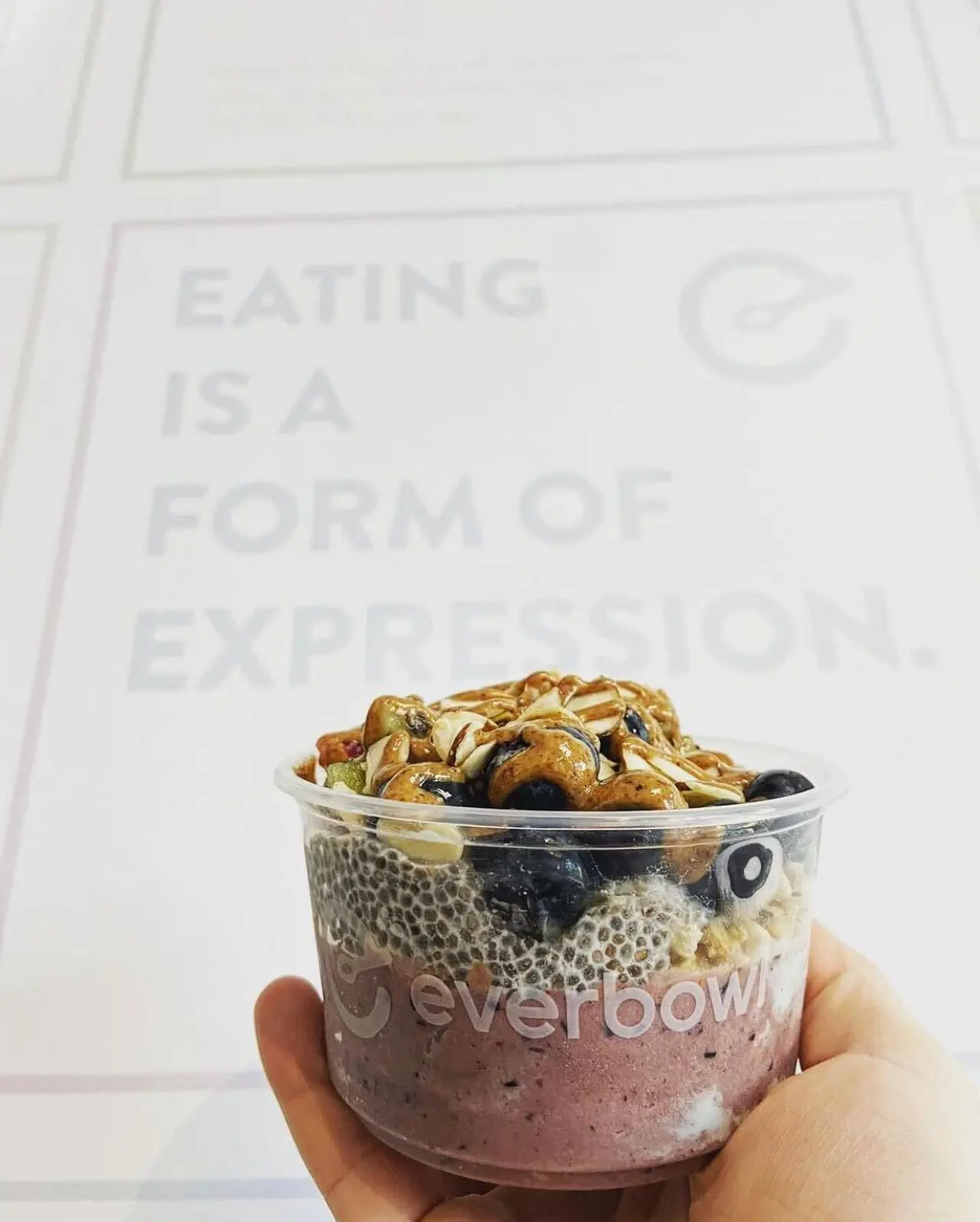 Hand holding clear bowl of acai base topped with granola and fruit against white wall with text