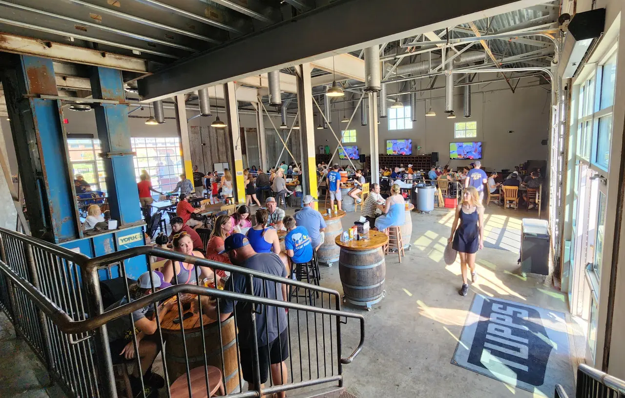 Large room with people, beer barrels, tables, and metal fixtures