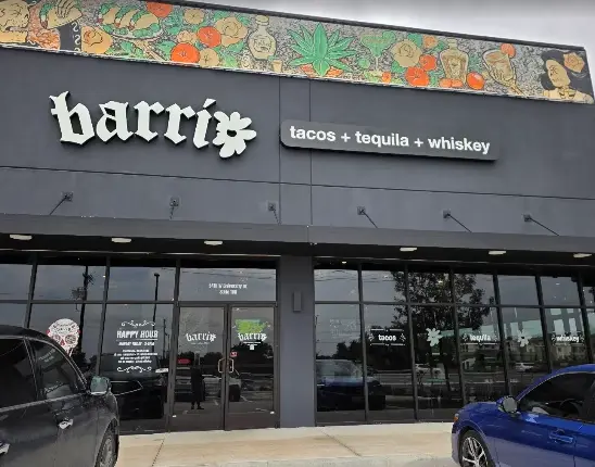 Dark storefront with white signage reading "barrio" and "tacos + tequila + whiskey," colorful floral border above, parked cars in front