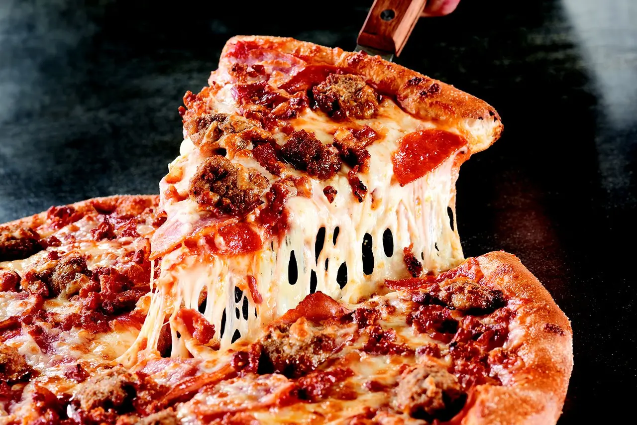 Slice of pizza with melted cheese and toppings being lifted from pie on dark surface
