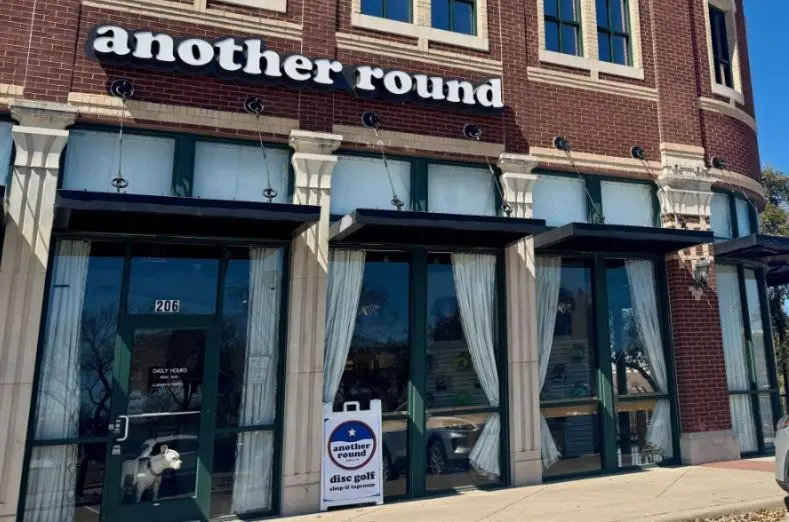 Storefront with large windows, white columns, and "another round" signage on red brick building facade