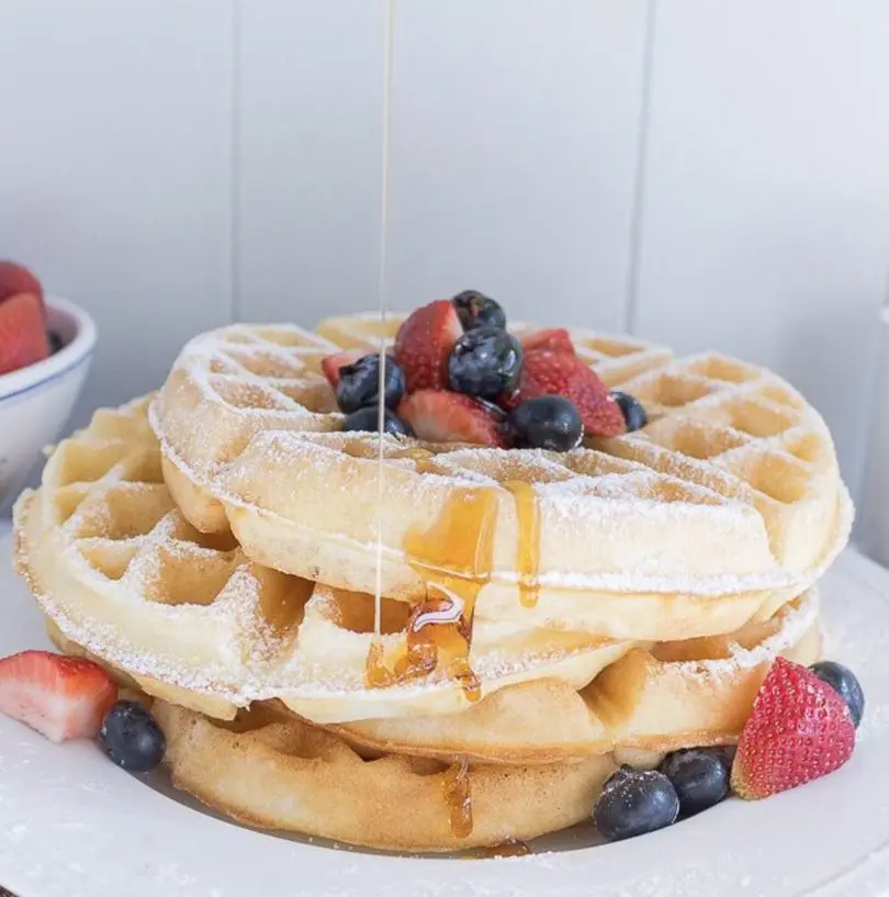 Stack of Belgian waffles dusted with powdered sugar, topped with fresh strawberries and blueberries, drizzled with syrup