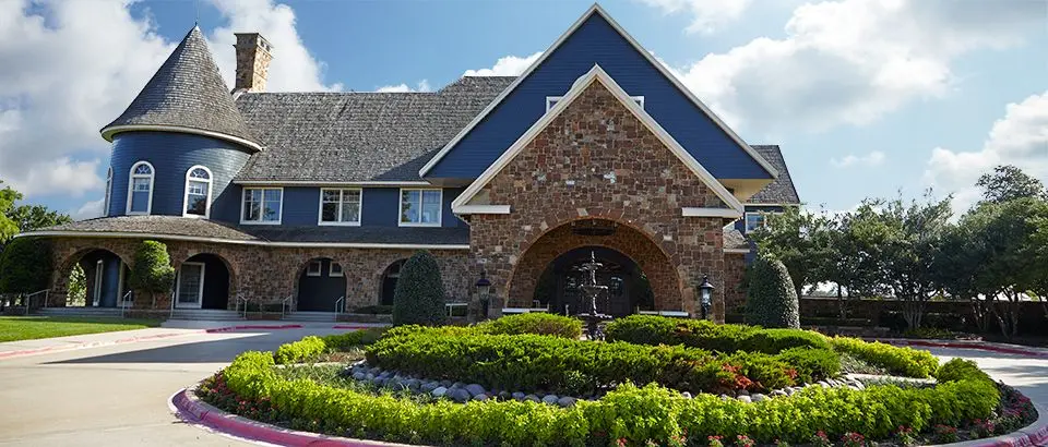 Large brick and stone country club building with turrets and arched entrance, manicured landscaping with purple mulch in foreground