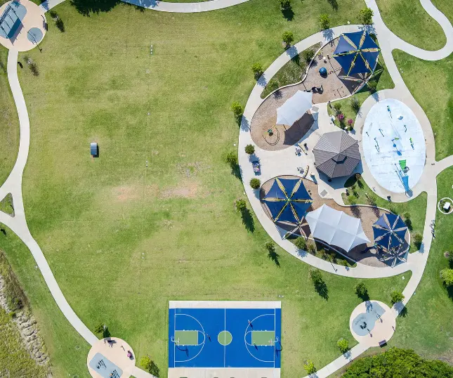 Aerial view of neighborhood park with sports court, shaded seating areas, open grass field, and walking paths
