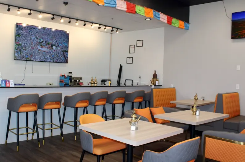 Modern restaurant interior with orange and gray seating, white tables, bar counter with stools, and colorful prayer flags above