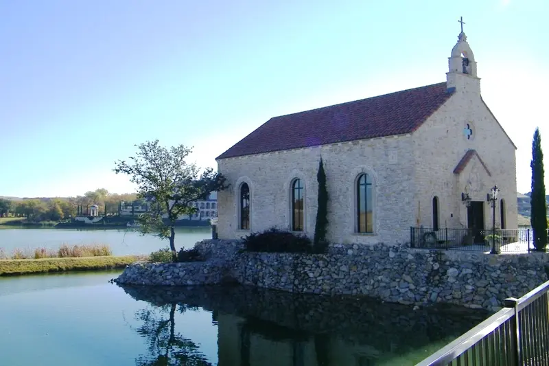 White stone chapel with dark roof and cross on hilltop, surrounded by water and cypress trees