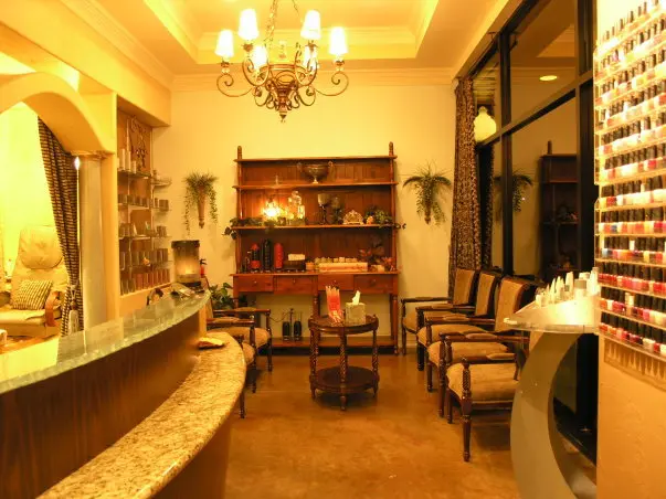 Spa reception area with curved counter, wooden chairs, shelving with products, chandelier, and warm golden lighting