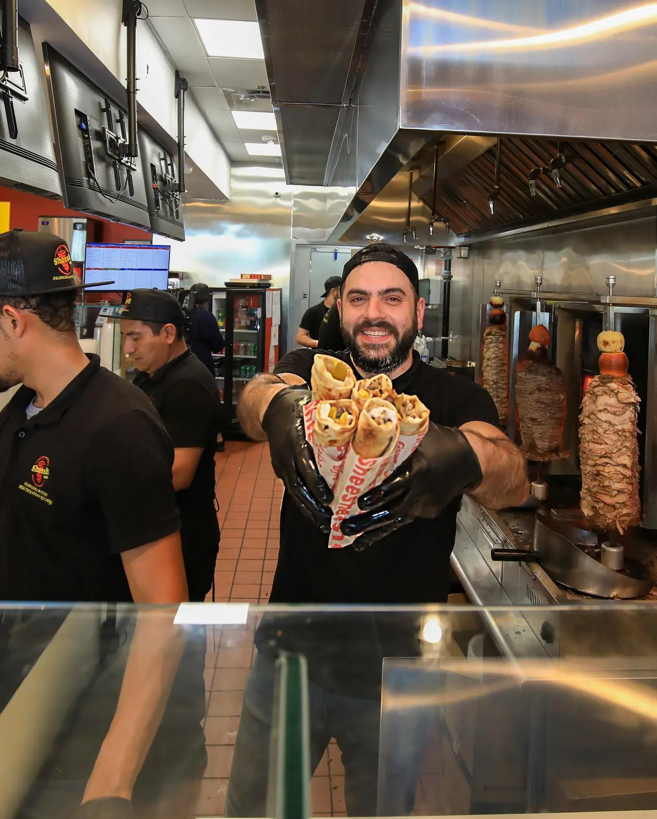 Chef holding wrapped shawarma in front of counter with vertical meat rotisseries in kitchen