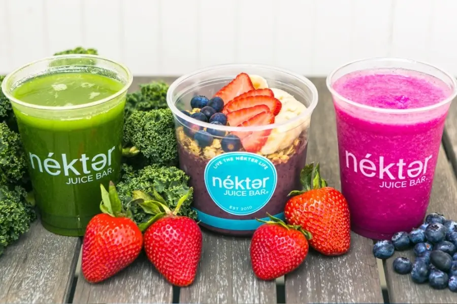 Three juice drinks in clear cups: green juice with kale, pink berry smoothie, and acai bowl with berries and granola, surrounded by fresh strawberries and
