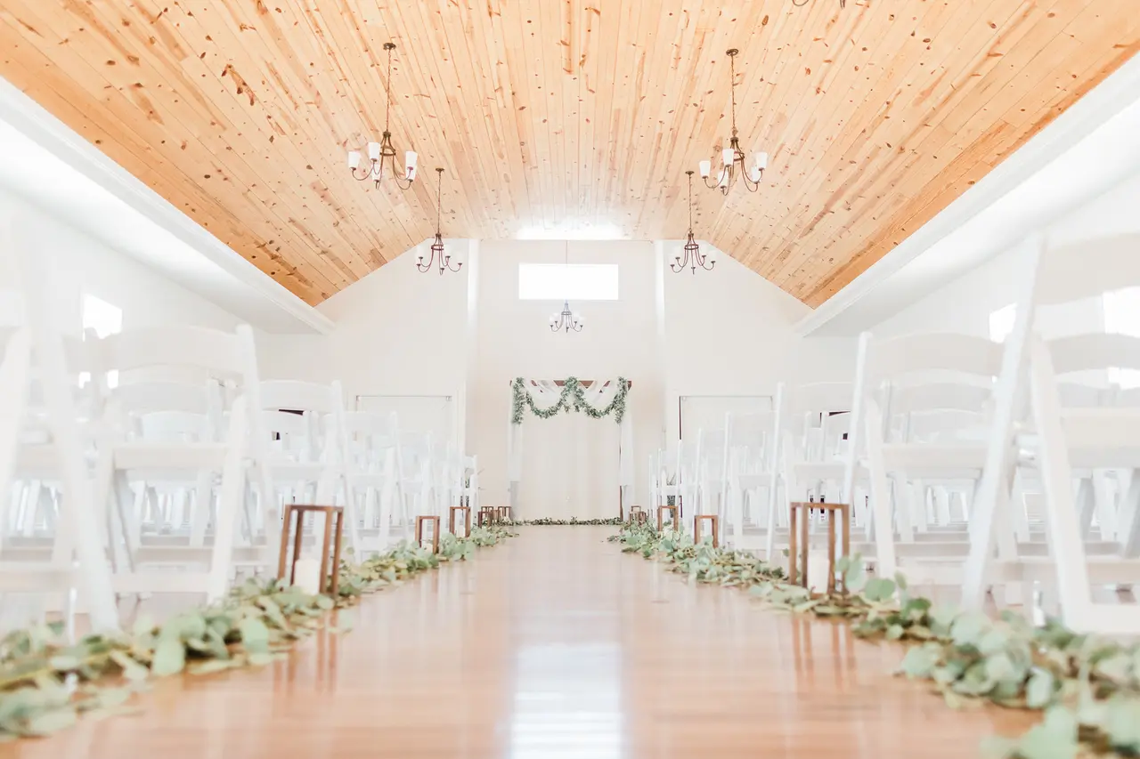 Bright, modern indoor event space with wooden vaulted ceiling, white walls, pendant lights, and floral garland lining the aisle