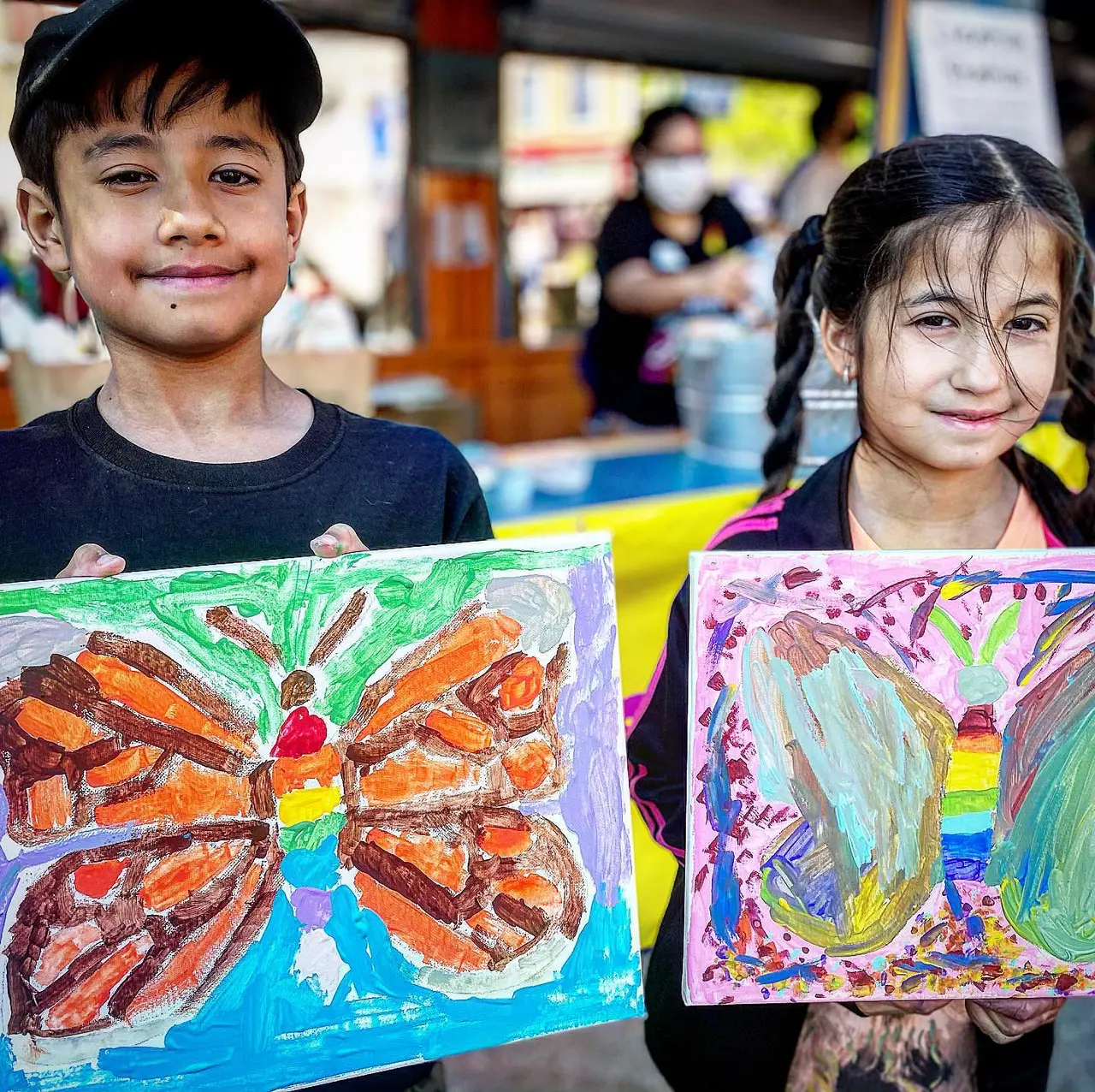 Two children holding colorful painted artwork featuring a butterfly and landscape scene in an art studio
