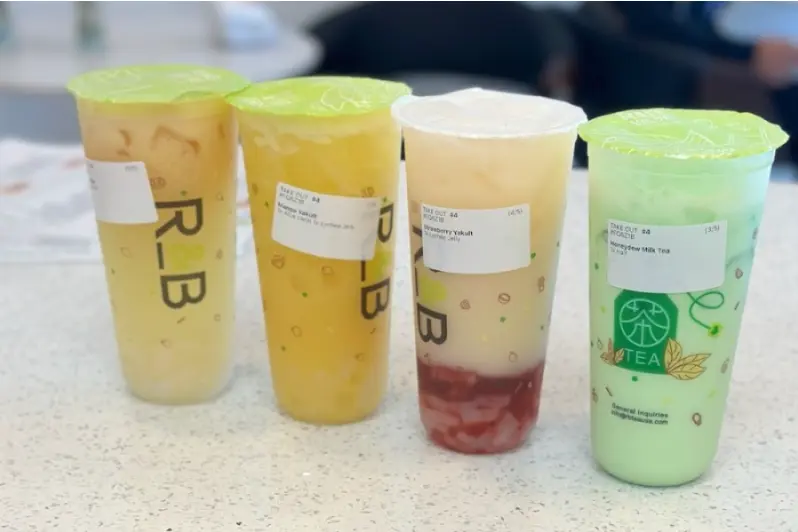 Four bubble tea drinks in clear cups with plastic lids, ranging from pale yellow to green, displayed on a white counter
