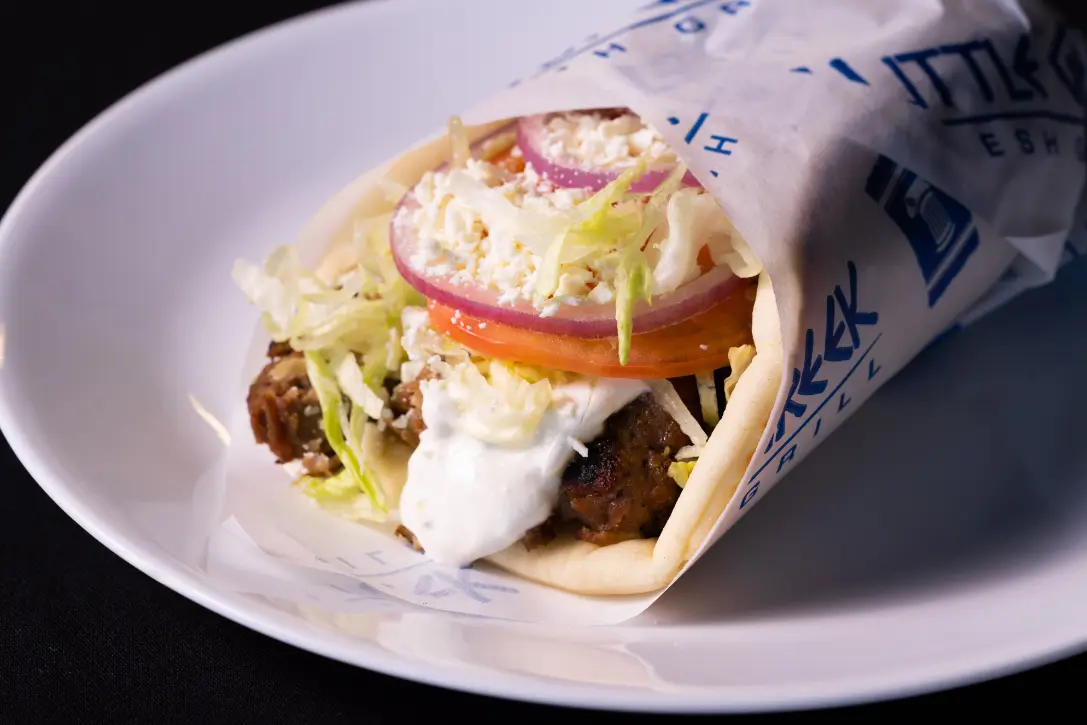 Gyro wrap filled with seasoned meat, tzatziki sauce, sliced tomatoes, onions, and lettuce on white plate