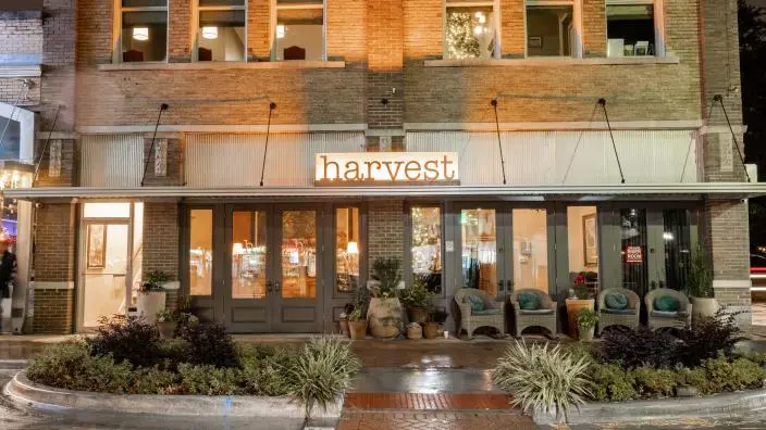 Storefront of farm-to-table restaurant with illuminated signage, large windows, brick facade, and potted plants flanking the entrance