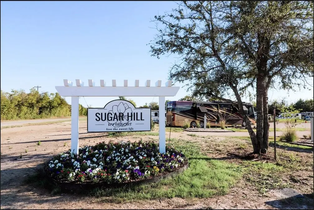 White entrance sign reading "Sugar Hill" with flowering planters, large tree, and RV parked in background under clear sky