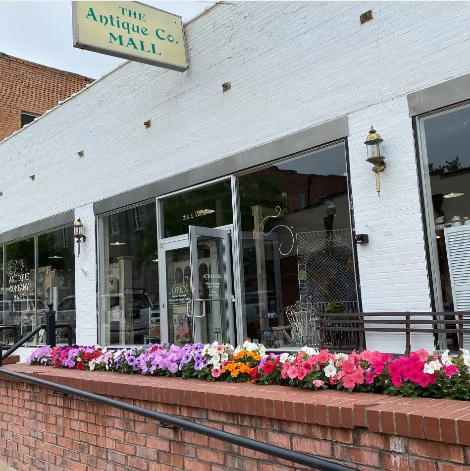 White storefront with large windows displaying antique goods, colorful flowers in window box planters