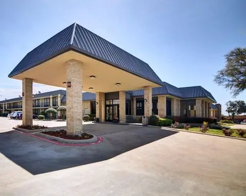 Modern hotel building with peaked metal roof, stone facade, covered entry portico, and curved driveway with landscaping under clear sky