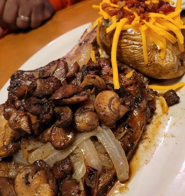 Grilled steak topped with sautéed mushrooms and onions, served with baked potato and cheese fries on white plate