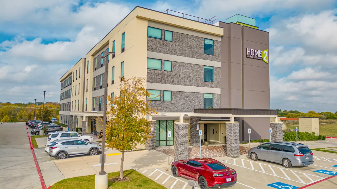 Modern four-story hotel building with stone and yellow facade, parking lot with several vehicles, young tree, and clear sky