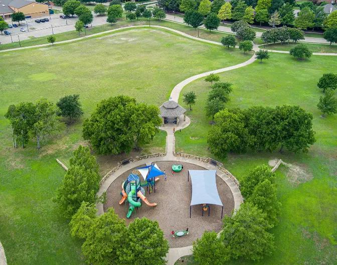 Aerial view of neighborhood park with playground equipment, shade structure, and curved walking path through grassy lawn dotted with trees
