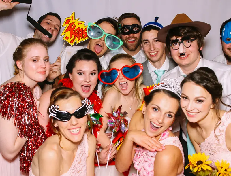 Group of people in formal attire posing together with colorful novelty props and oversized glasses at a photo booth