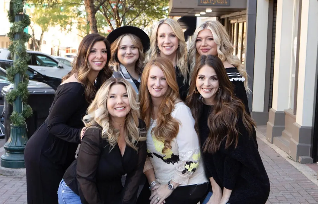 Group of seven women posing together on a downtown street, one wearing white and holding a bouquet, others in black clothing