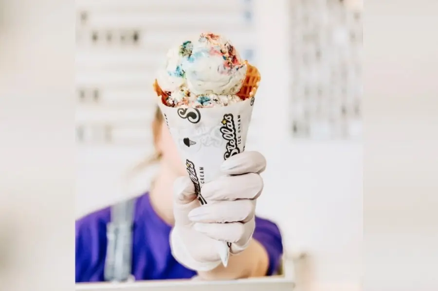 Person in blue shirt holding vanilla ice cream cone with colorful toppings and waffle cone wrapper visible