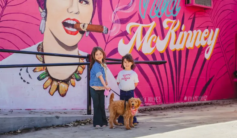 Two people and a brown dog standing in front of a colorful pop art mural with a woman's portrait and pink lettering