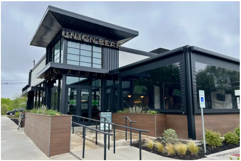 Modern brewery building with black metal and glass exterior, wood paneling, metal railing, and landscaping along the entrance