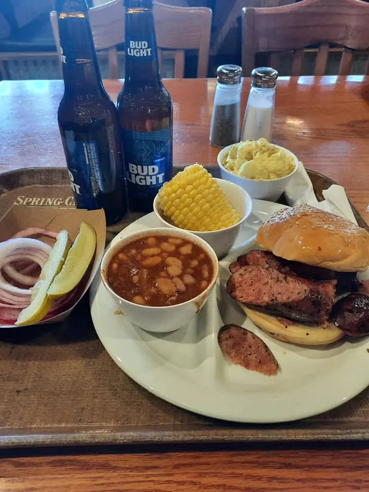 Plated BBQ meal with smoked meat sandwich, ribs, sausage, corn, beans, onions, and Bud Light beers on wooden table