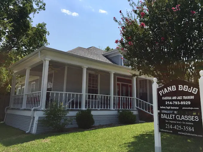 White cottage-style building with front porch columns and flowering vines, green lawn, and signage for Piano Dojo music lessons