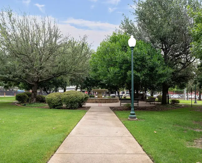 Concrete pathway lined with mature trees and green lawn leading toward a central plaza with a gazebo structure