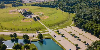 Aerial view of baseball and softball fields with parking area, pond, and surrounding green space and trees