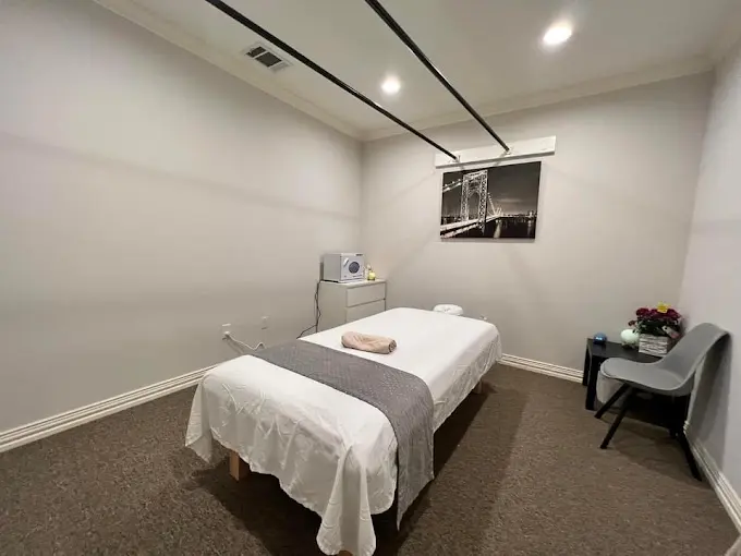 Minimalist massage treatment room with two beds draped in white linens, small side table, and mounted television