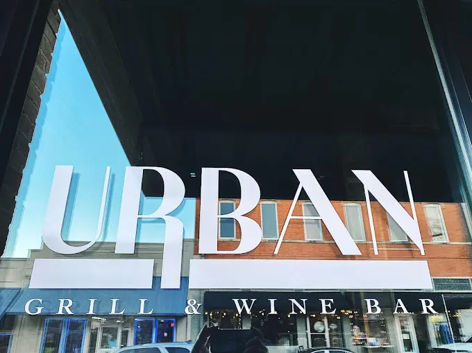 Large illuminated sign reading "Urban" mounted on storefront with "Grill & Wine Bar" signage below, viewed from street level against blue sky